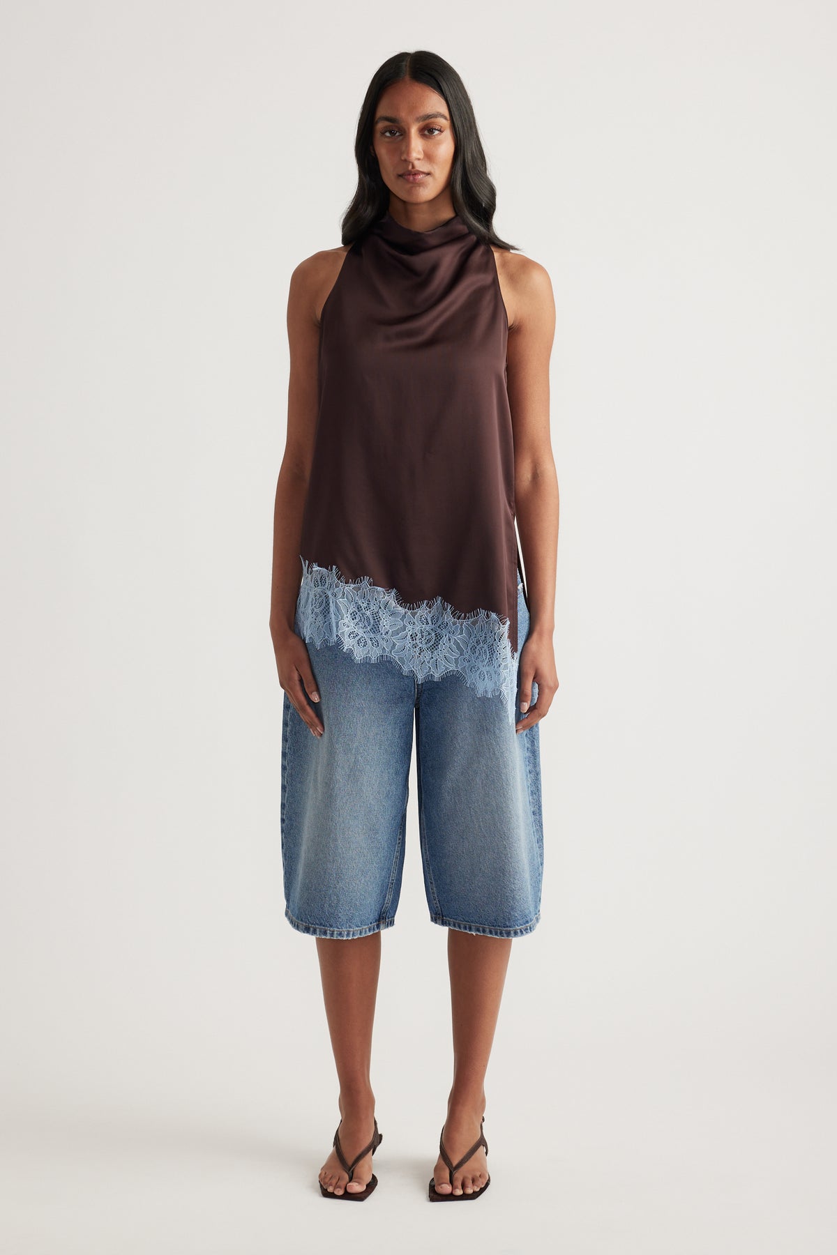 Woman wearing a chocolate brown sleeveless cowl neck top with lace trim and cropped blue wide leg jeans