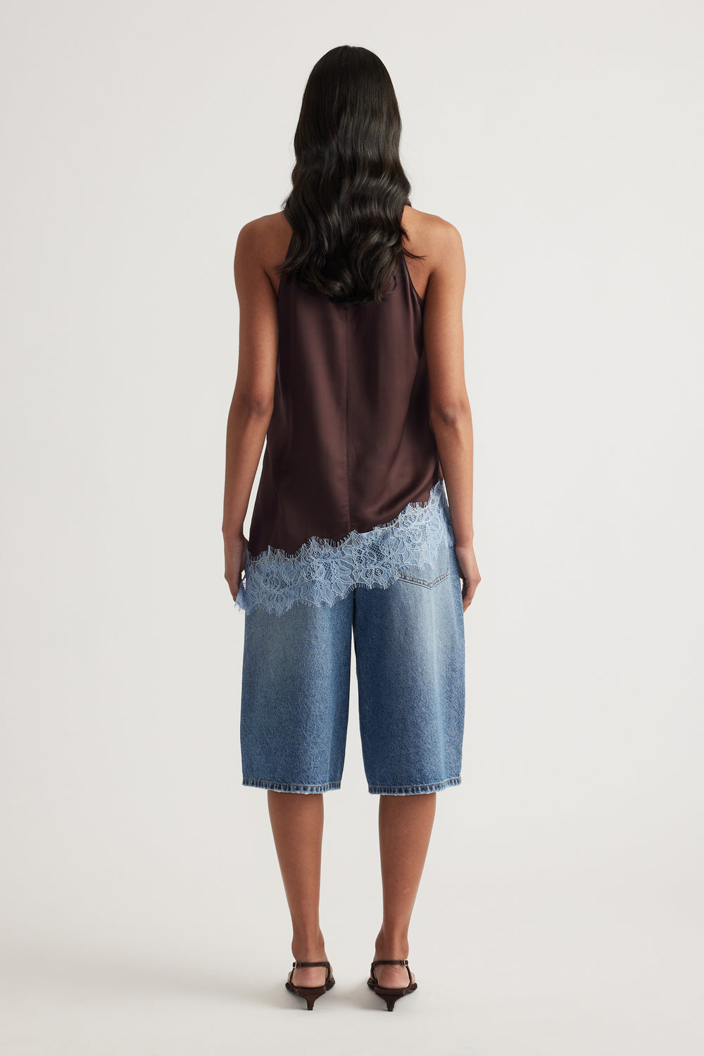 Back view of a woman in a brown sleeveless lace trim top, denim shorts and black heels