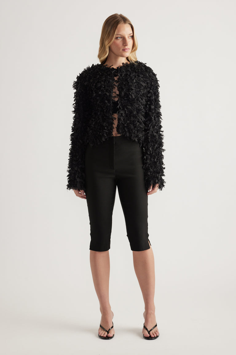 Model wearing a black textured jacket with long sleeves and a round neckline, styled with black pants and sandals