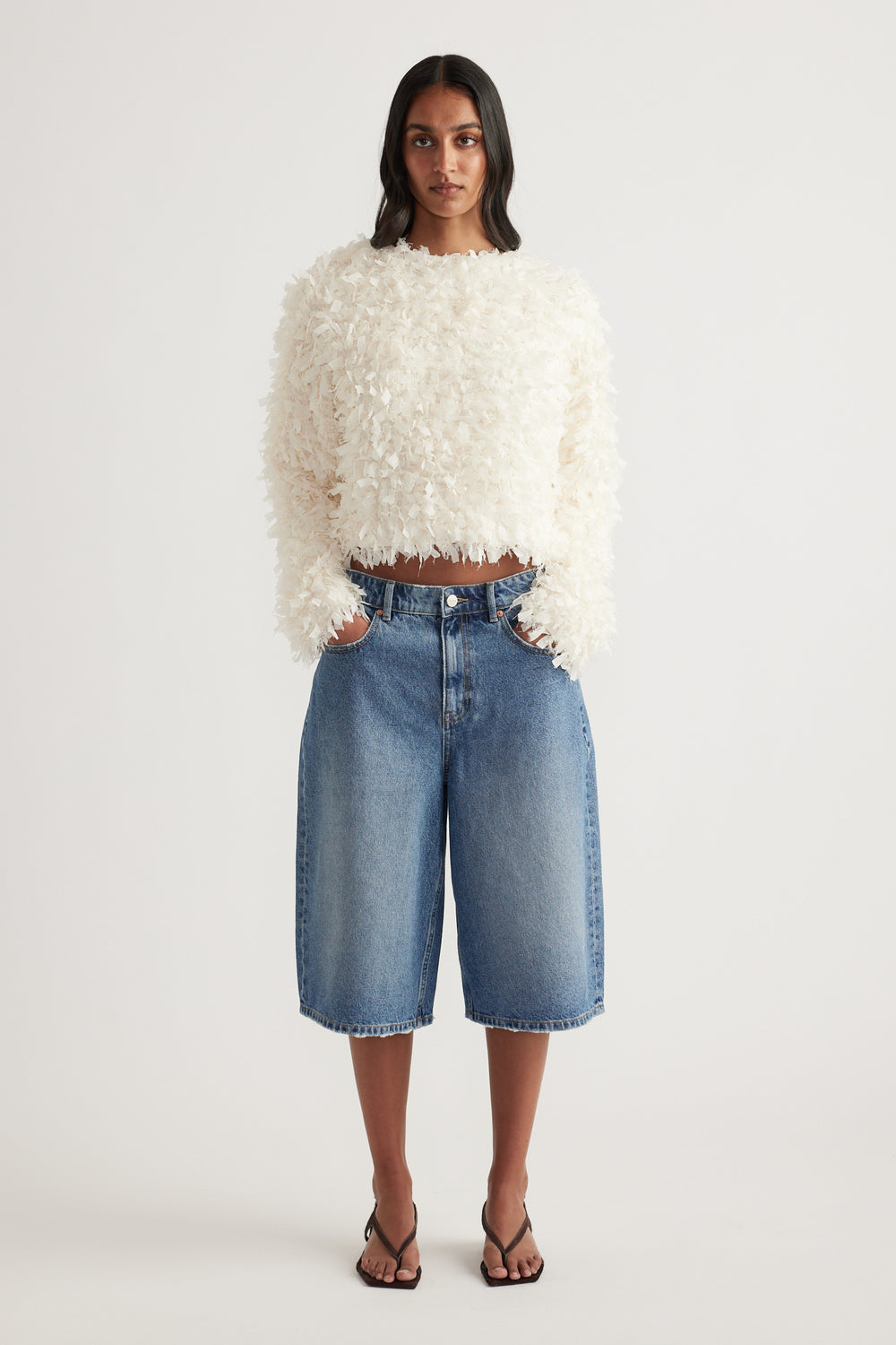 Model wearing a cream textured long sleeve jacket with denim bermuda shorts and strappy sandals