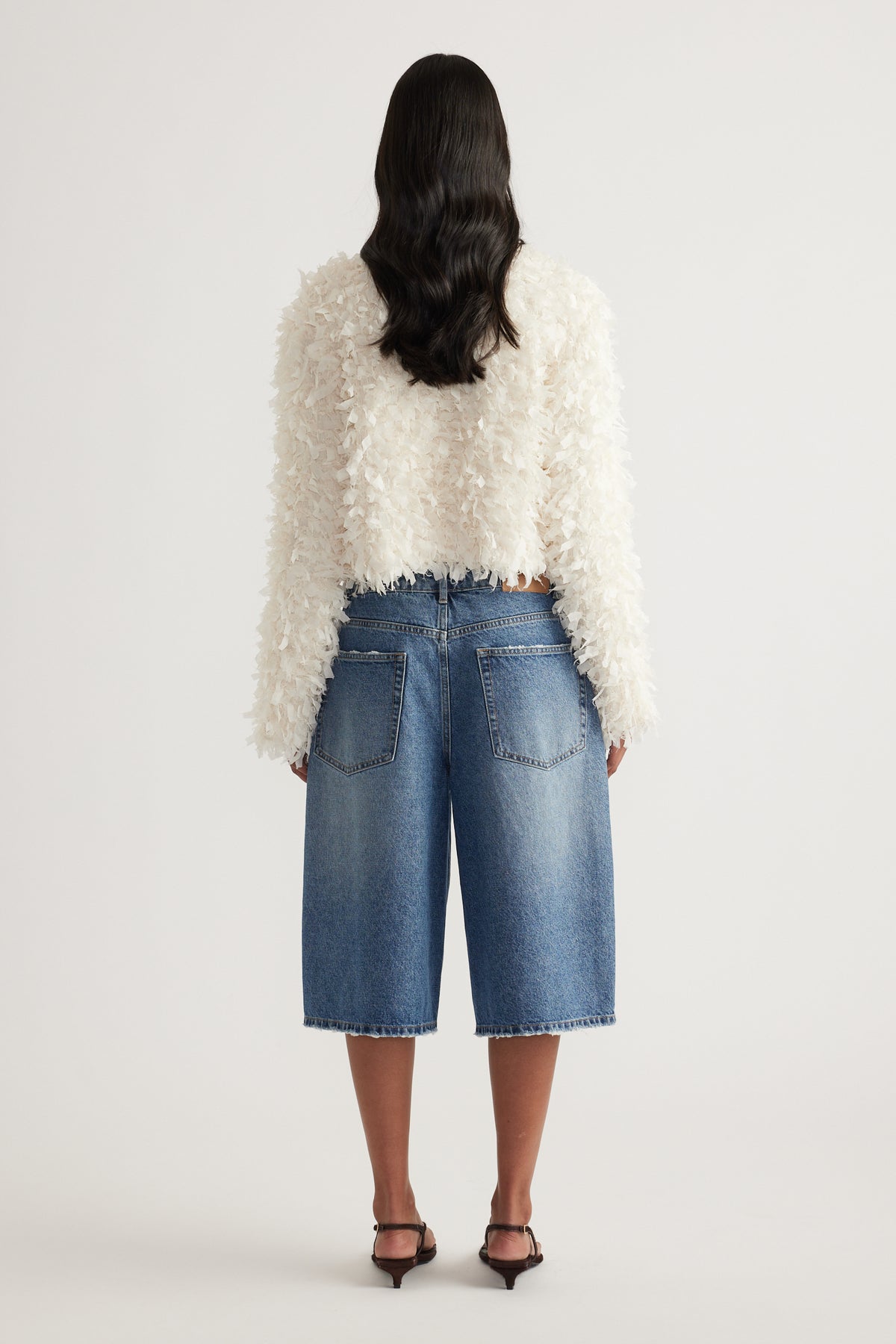 Back view of woman in a cream textured jacket, knee length denim shorts and black heels