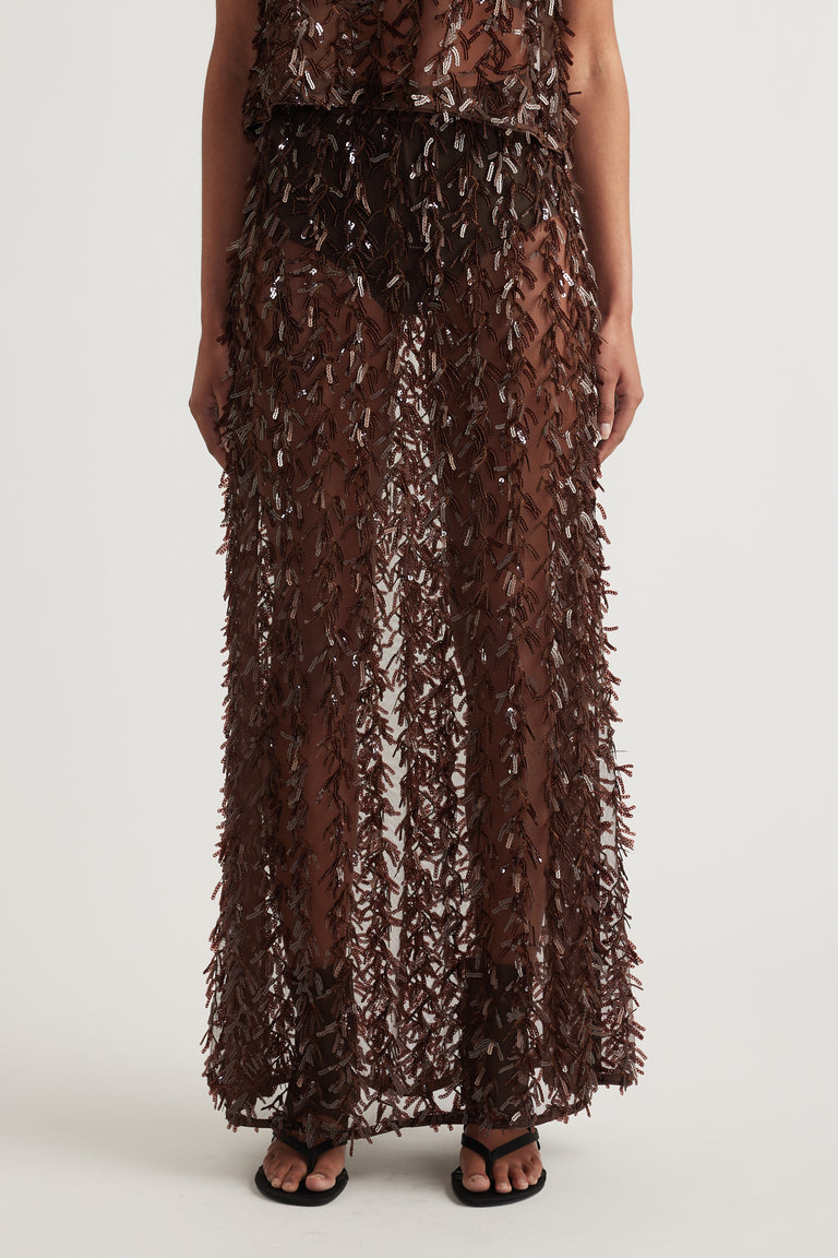 Woman wearing a brown sequin maxi skirt with sheer fringed texture and black sandals