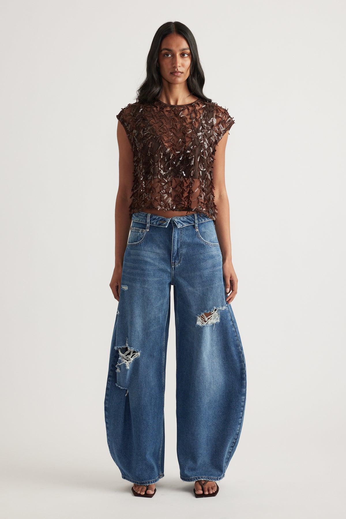 Woman in a brown distressed sleeveless top with relaxed blue ripped barrel leg jeans and black sandals