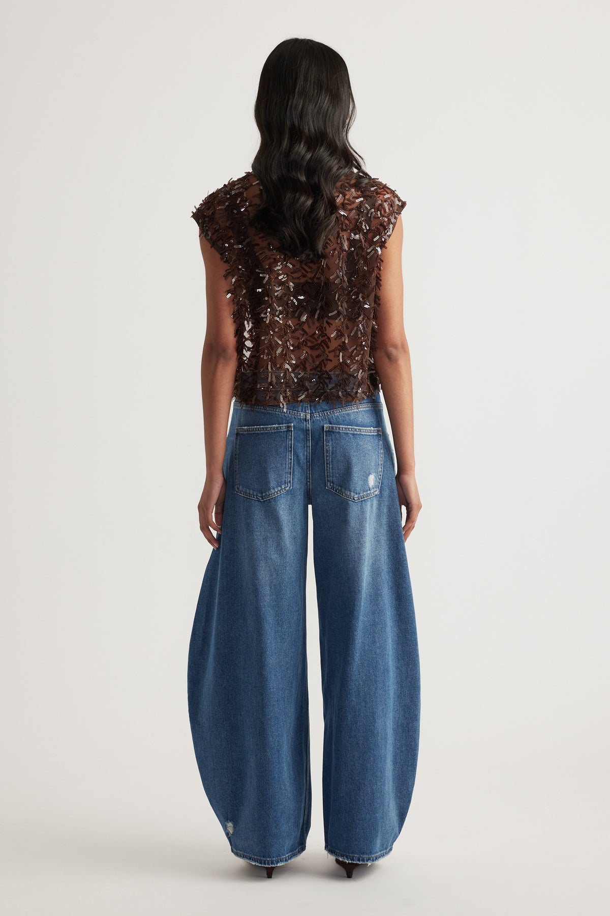 Woman seen from behind wearing a brown embellished sleeveless top and relaxed blue barrel leg jeans
