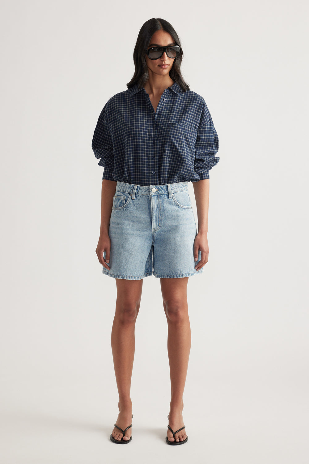 Woman wearing oversized navy check shirt with rolled sleeves paired with light wash denim shorts and black sandals