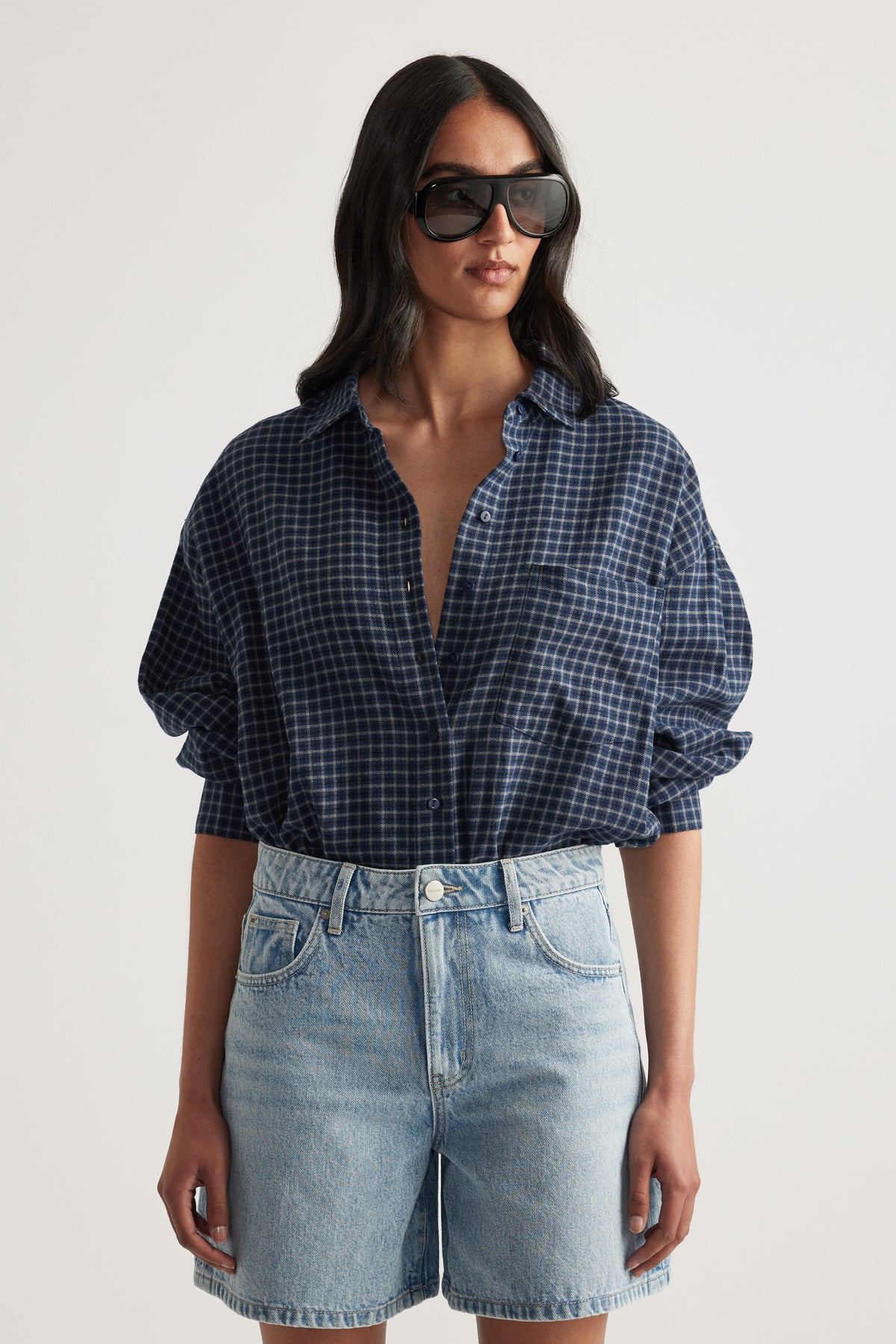 Woman wearing oversized navy checkered flannel shirt paired with light wash high-rise denim shorts and sunglasses