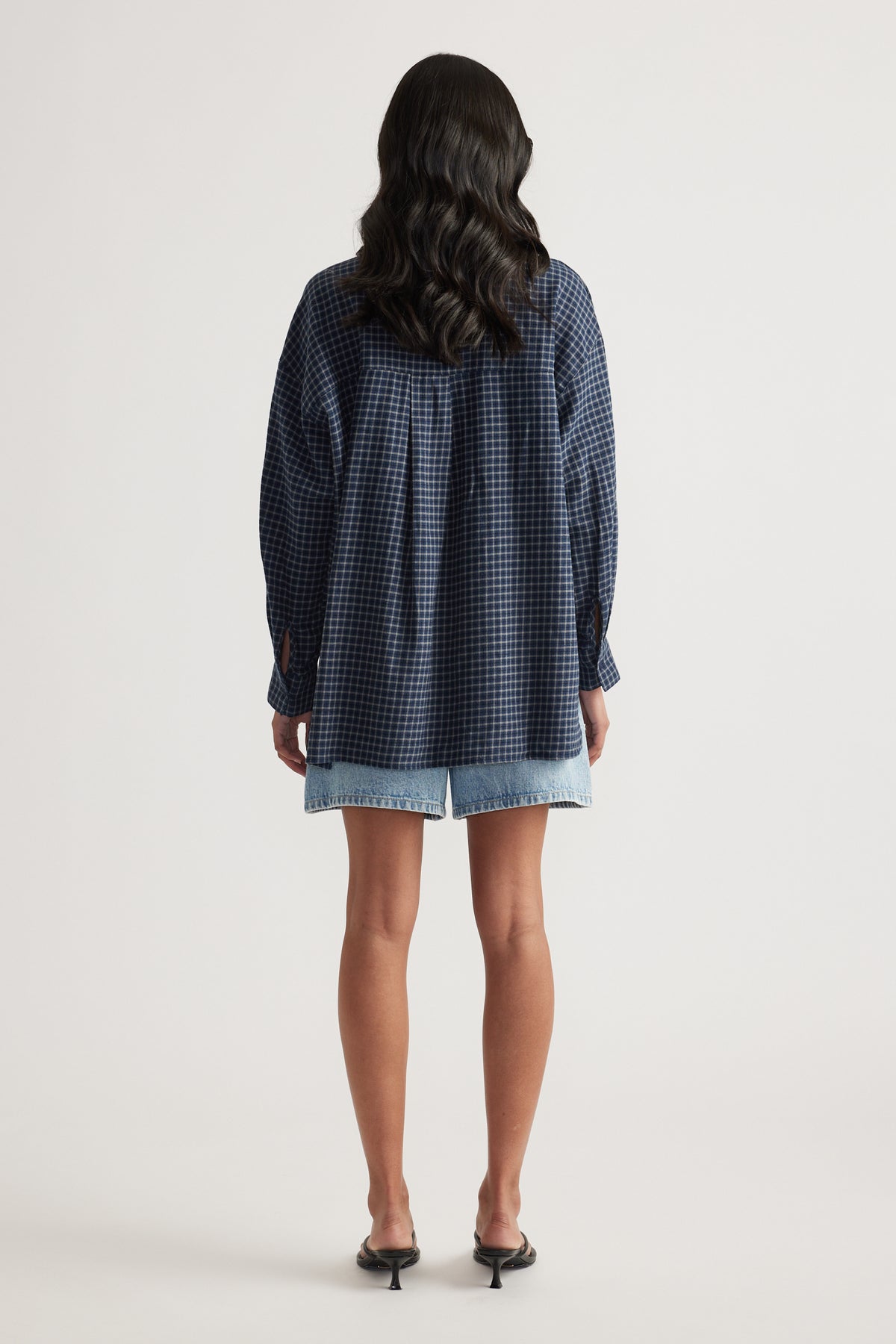 Rear view of a woman wearing an oversized navy check flannel shirt with light blue denim shorts and black heels