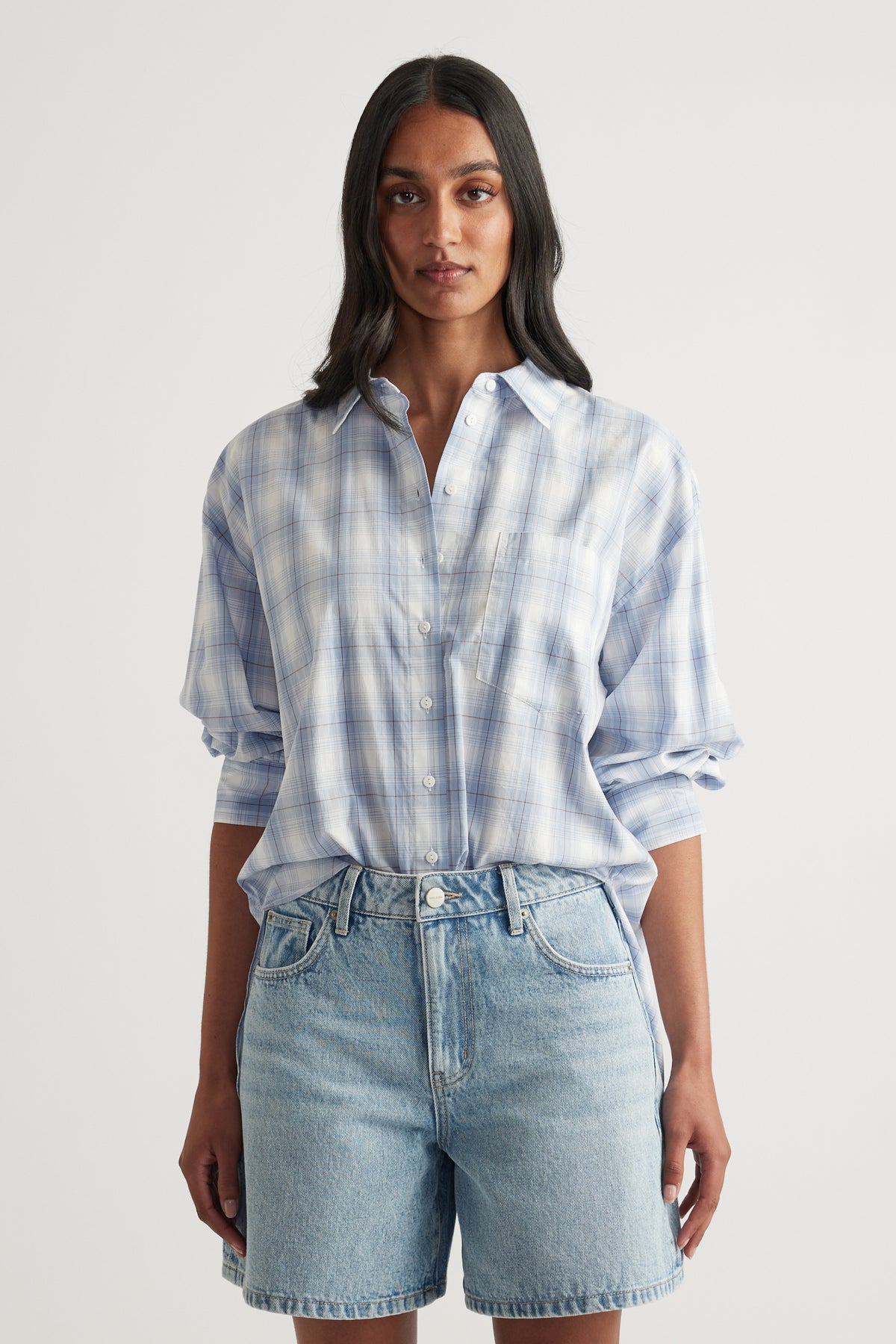 Woman wearing an oversized blue and white plaid shirt with button front and rolled sleeves, styled with denim shorts