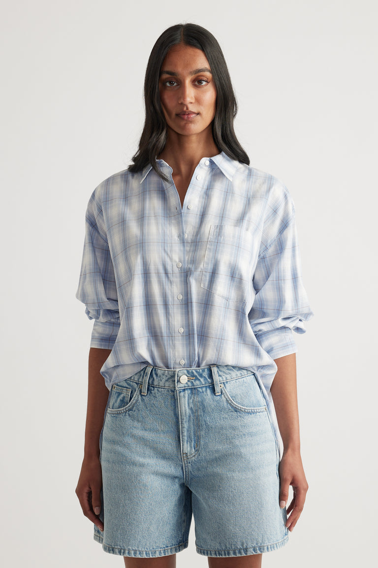 Woman wearing an oversized blue and white plaid shirt with button front and rolled sleeves, styled with denim shorts