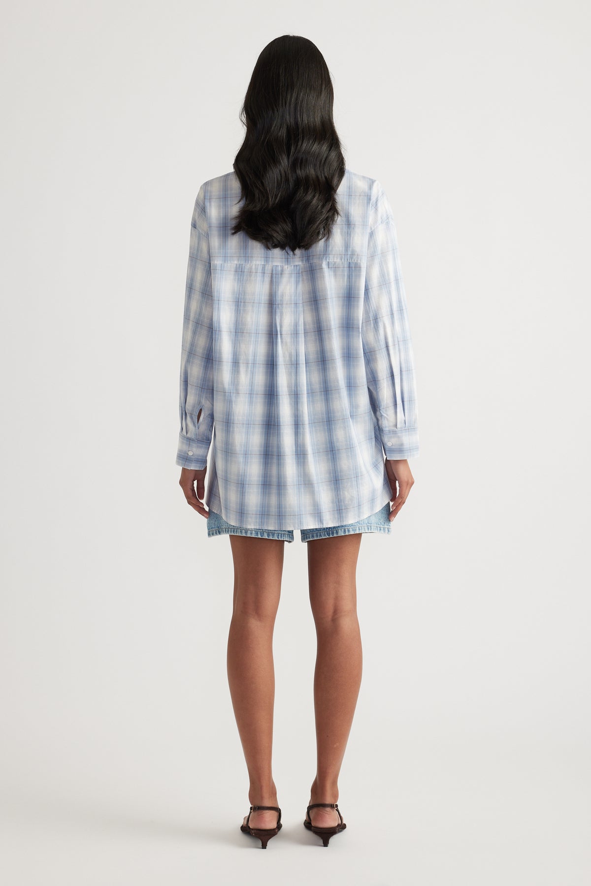 Woman viewed from behind wearing an oversized blue and white checked shirt with long sleeves and denim shorts
