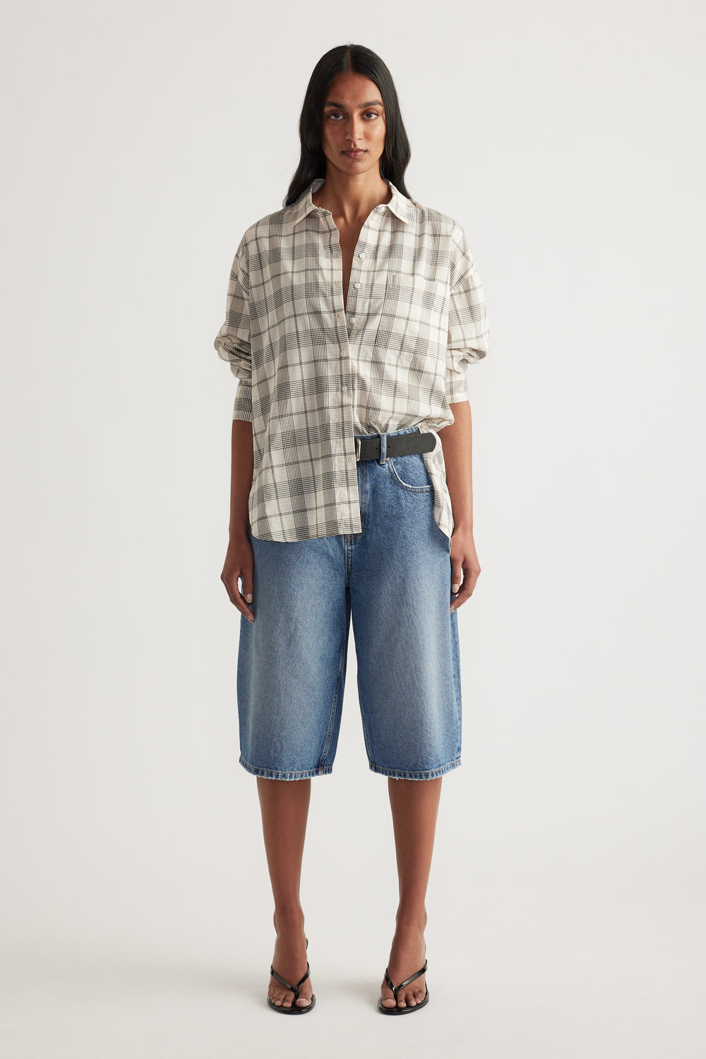 Woman wearing an oversized beige plaid button front shirt with rolled sleeves and knee length denim shorts