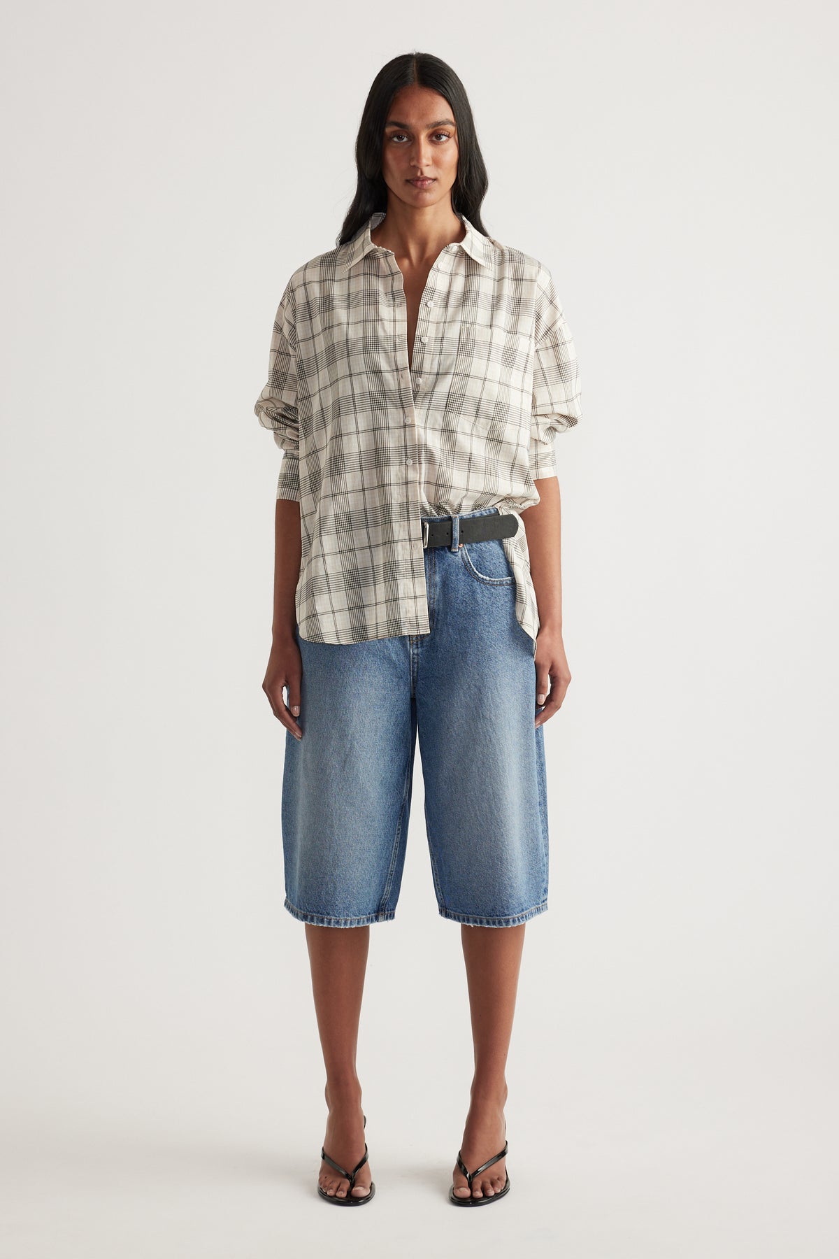 Woman wearing an oversized beige plaid button front shirt with rolled sleeves and knee length denim shorts
