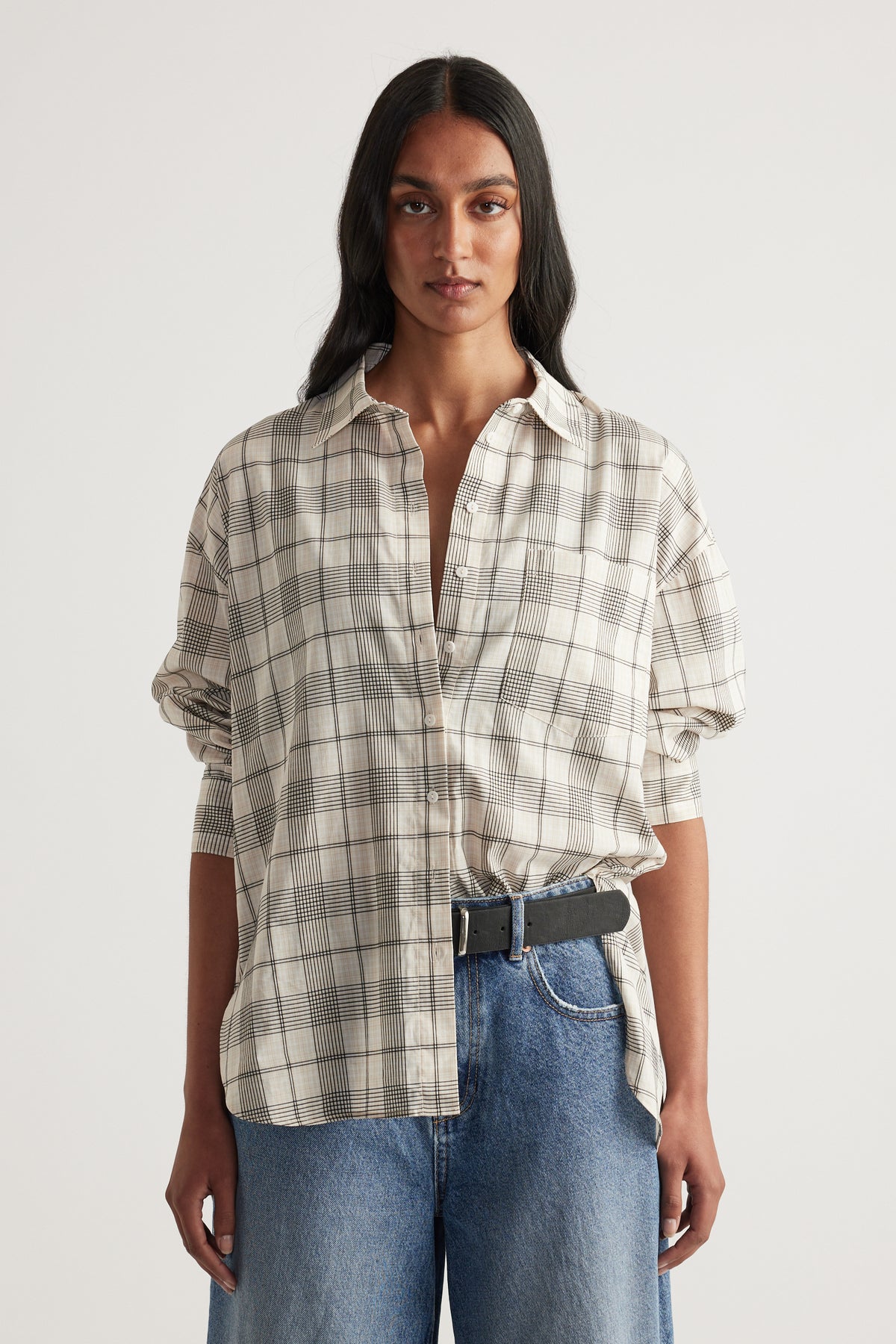Woman wearing an oversized beige plaid shirt with chest pocket and rolled sleeves, blue jeans and a belt