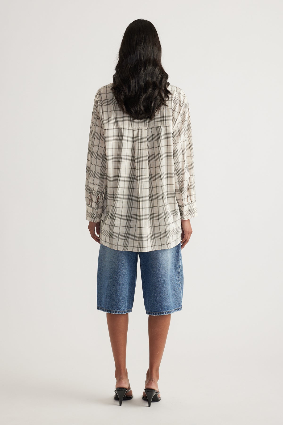 Woman viewed from behind wearing an oversized beige plaid shirt with long sleeves and denim shorts