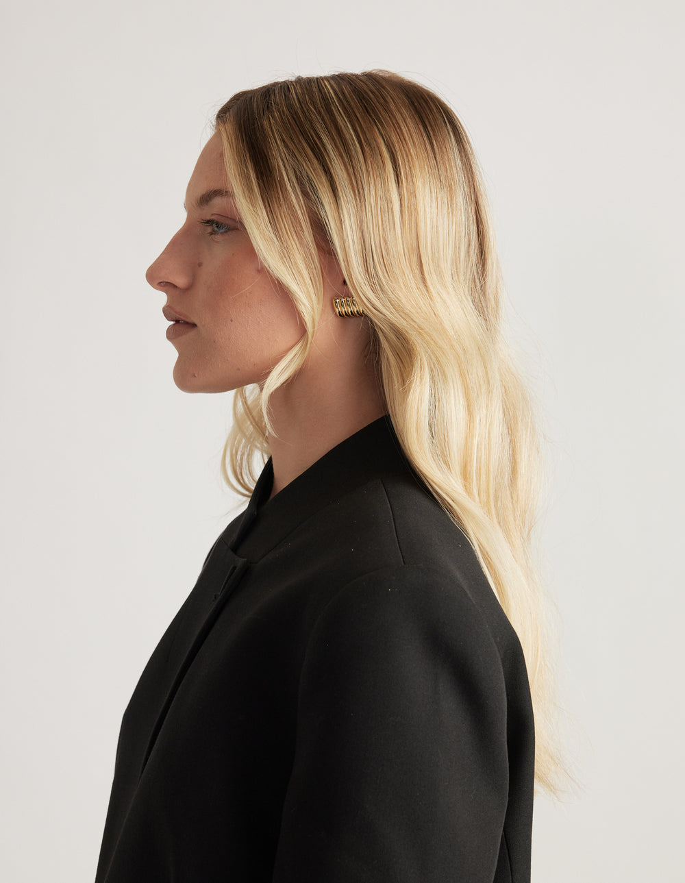 Woman in profile wearing a black blazer with a gold ribbed stud earring and long blonde hair