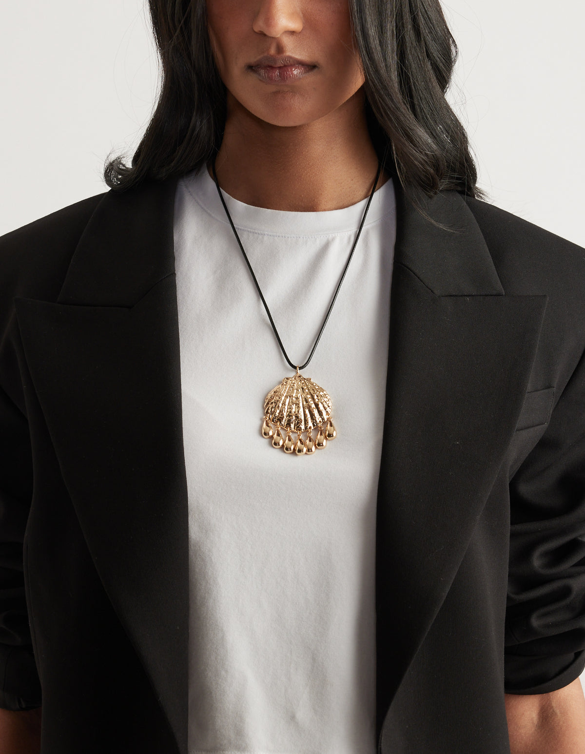 Woman wearing a gold textured shell pendant necklace over a white tee and black blazer