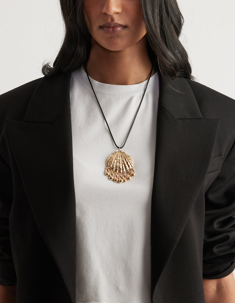 Woman wearing a gold textured shell pendant necklace over a white tee and black blazer