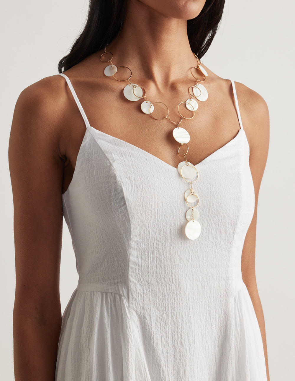 Layered gold lariat necklace with pearlescent shells and tiered hoops worn over a white camisole dress