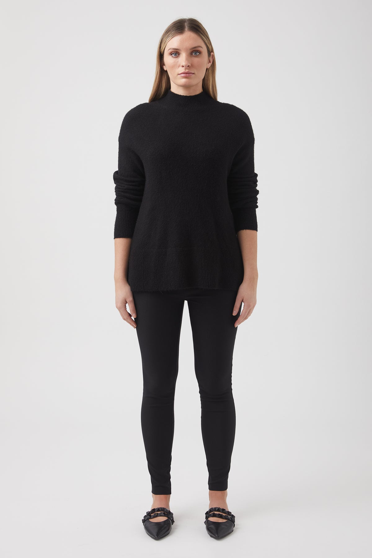 Woman wearing a black high-neck knit sweater paired with black slim pants and black pointed flats against a plain background