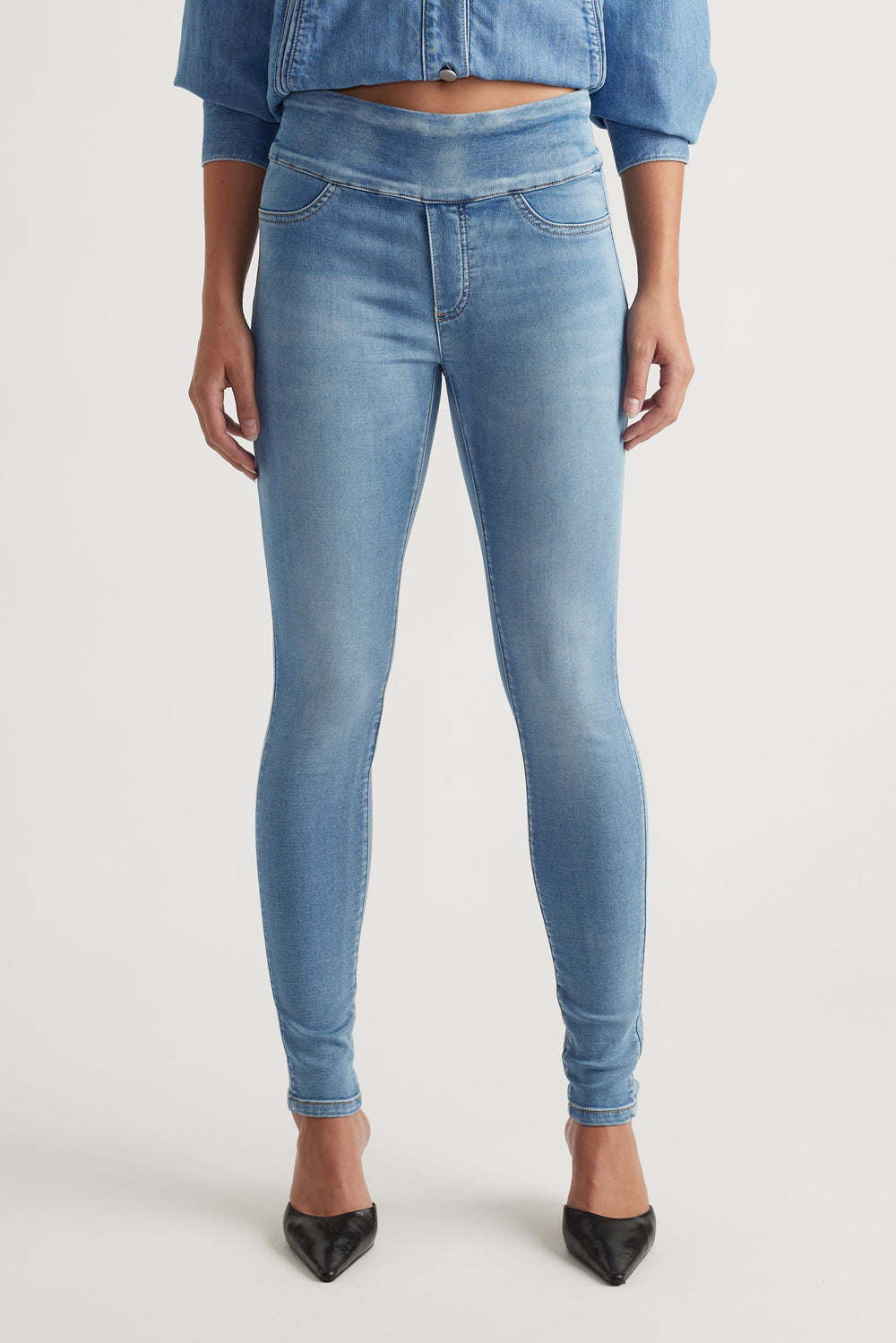 Front view of woman wearing light blue mid rise skinny jeans paired with black pointed heels and a cropped denim jacket