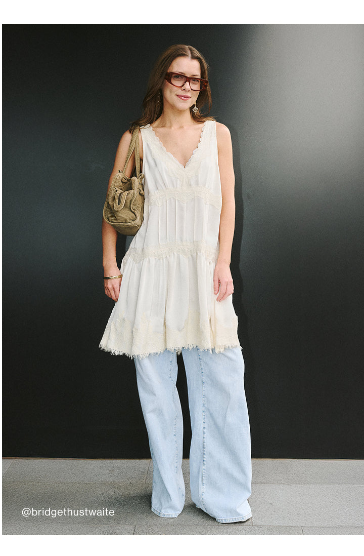 A woman modeling a sleeveless cream-colored tiered tunic top with light-wash wide-leg jeans and a tan shoulder bag, standing against a dark gray wall.
