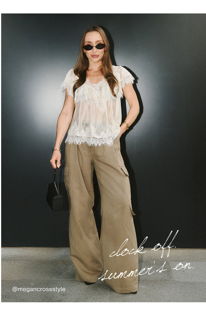 A woman models a white sheer ruffled short-sleeve top paired with wide-leg khaki cargo pants, styled with black sunglasses and a small black handbag against a dark gray studio background.