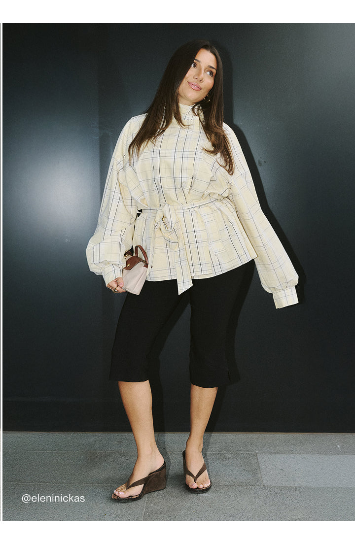 A woman modeling a cream and yellow plaid long-sleeve belted blouse with black cropped pants and wedge sandals, standing against a dark gray wall.