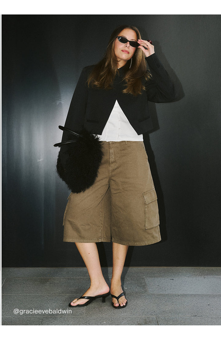 A woman models a black cropped jacket, white top, brown cargo shorts, and black sandals against a dark studio background.