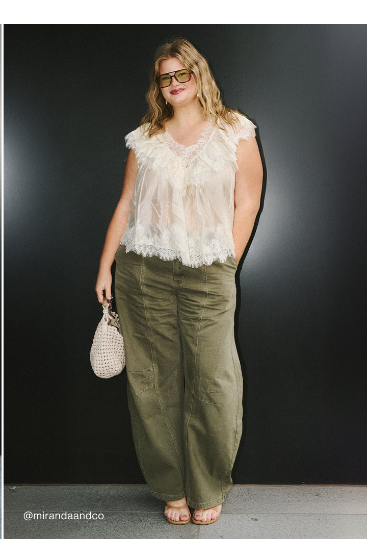 A woman modeling a white ruffled sleeveless top and olive green wide-leg pants, holding a small woven beige handbag against a dark studio background.