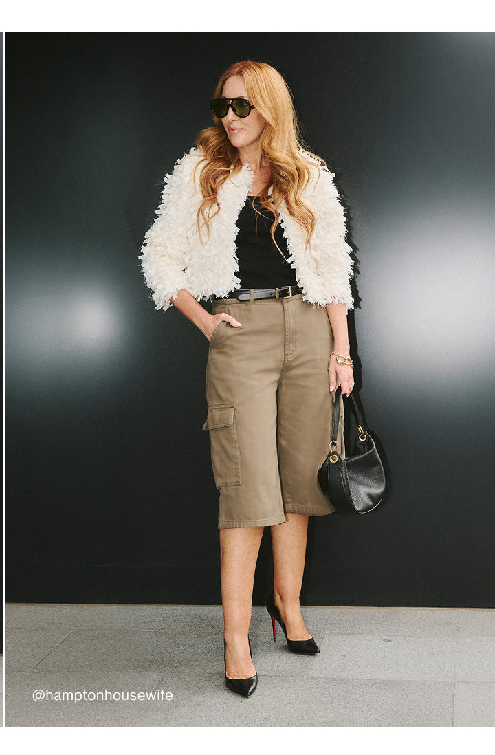 A woman models a fashion outfit consisting of a white fluffy cropped jacket, a black top, tan cargo midi skirt, black belt, black heels, and a black handbag against a dark gray studio background.