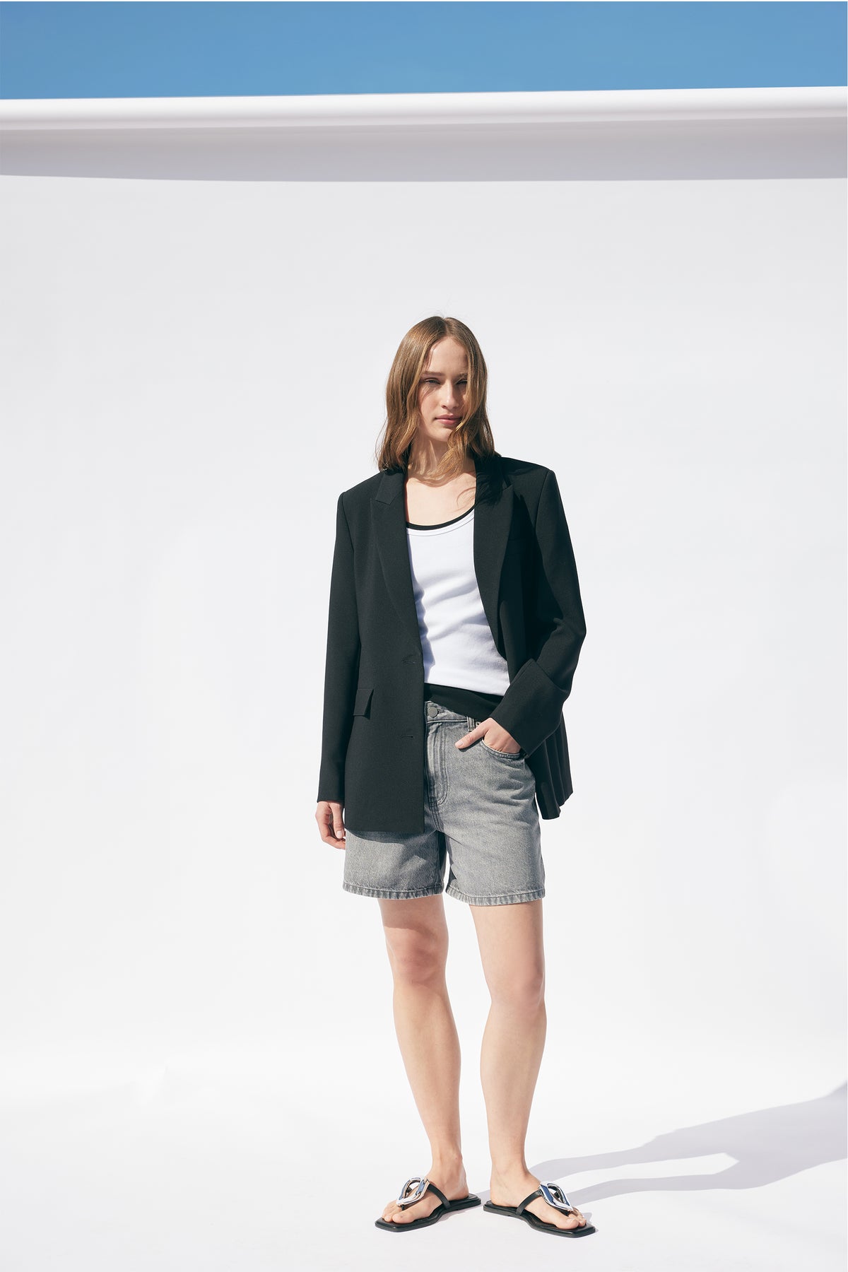 Woman wearing a black blazer over a white ribbed tank top with grey denim shorts and black sandals, standing outdoors