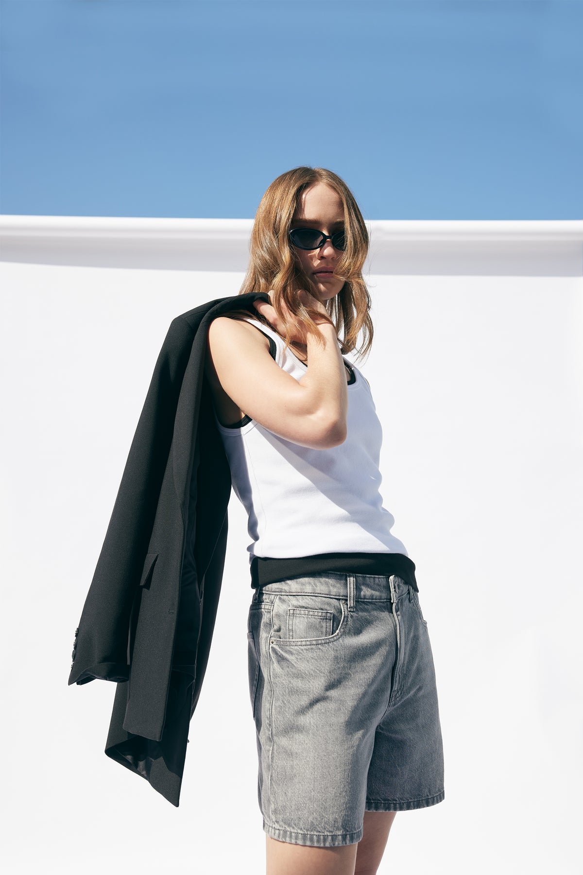 Woman wearing a white ribbed tank top paired with gray denim shorts and a black blazer draped over her shoulder