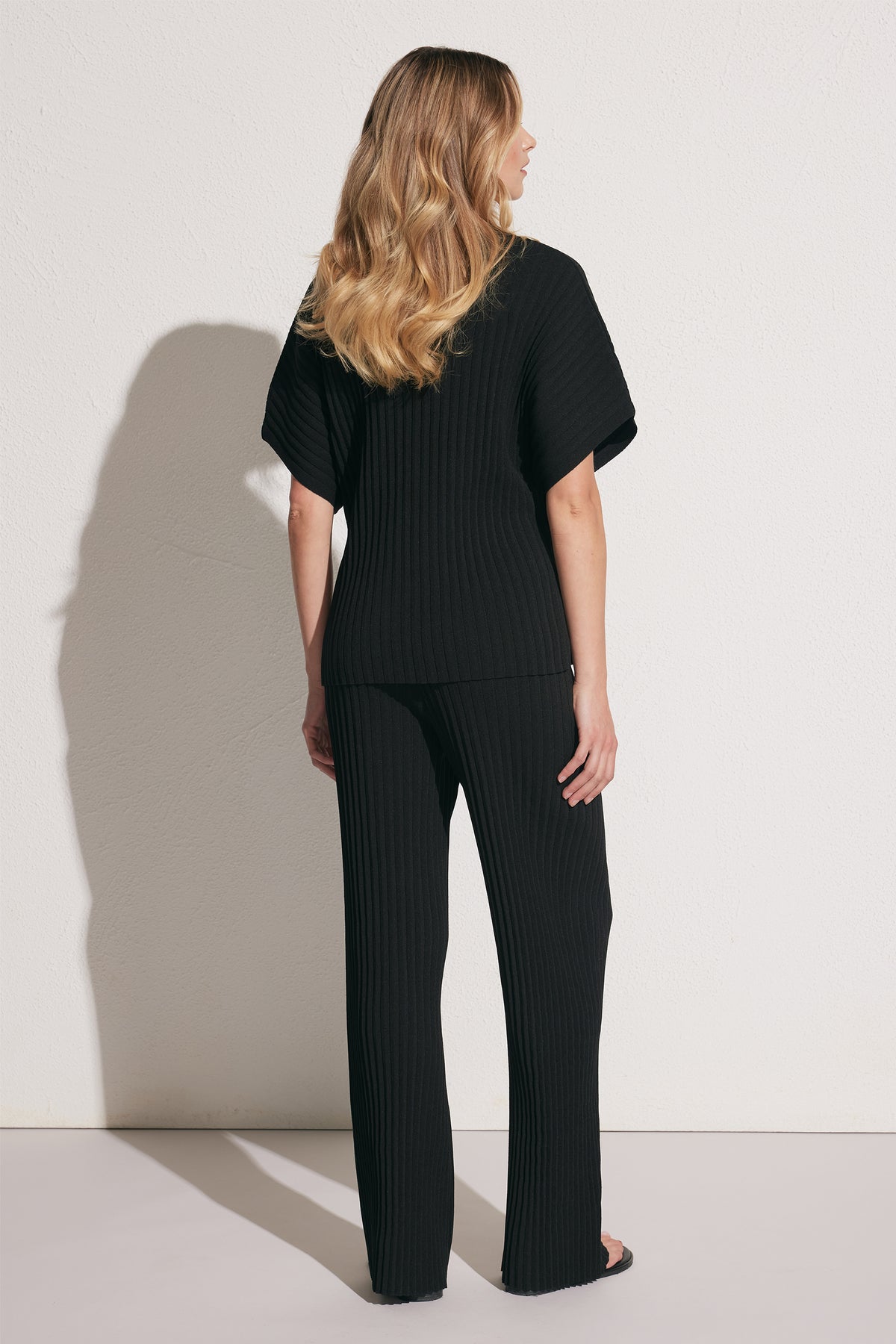 Woman wearing black ribbed knit pants and matching short-sleeve top, standing against a plain light wall, back view