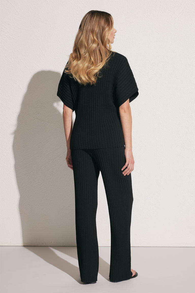 Woman wearing black ribbed knit pants and matching short-sleeve top, standing against a plain light wall, back view