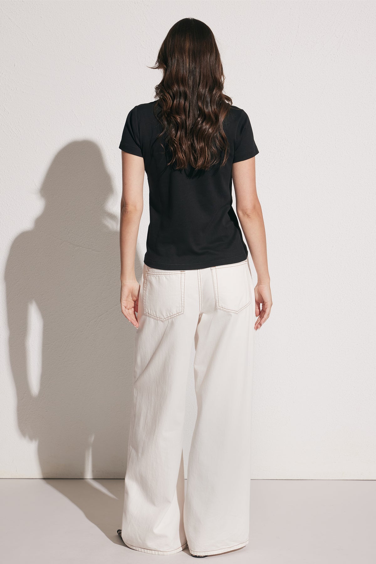 Woman wearing a fitted black crew neck tee paired with wide-leg white pants, back view against plain wall