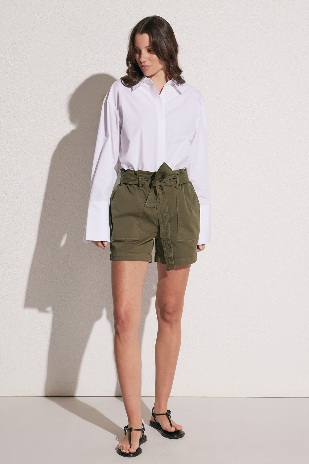 Woman wearing olive green high rise cargo shorts with a tie waist and patch pockets, styled with a white shirt and sandals