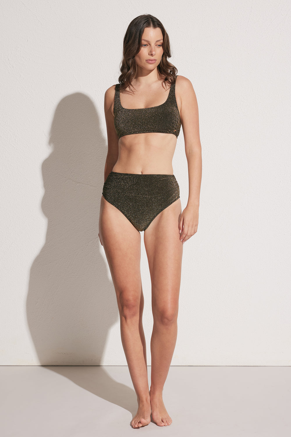 Woman wearing a shimmering gathered bikini bottom with a matching square-neck bikini top, standing barefoot against a light wall