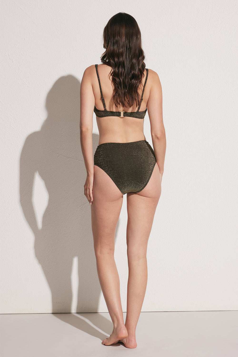 Woman wearing dark shimmer high-waisted bikini bottom with adjustable strap bikini top, back view against light wall