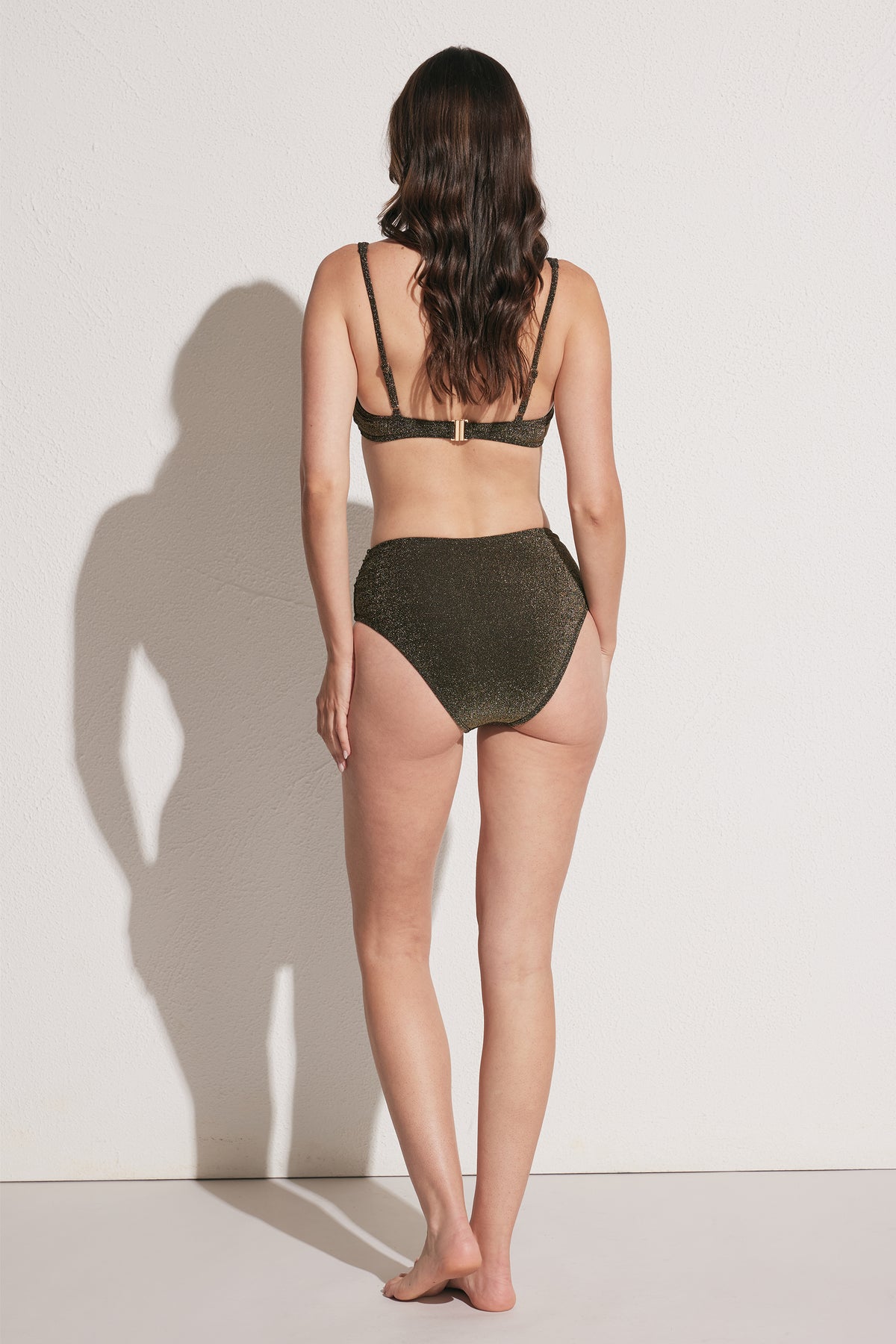Woman wearing dark shimmer high-waisted bikini bottom with adjustable strap bikini top, back view against light wall