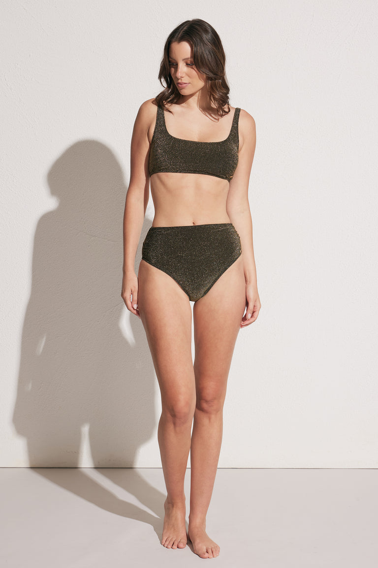Woman wearing shimmer textured black high-waisted bikini bottoms and square neck bikini top, standing against white wall
