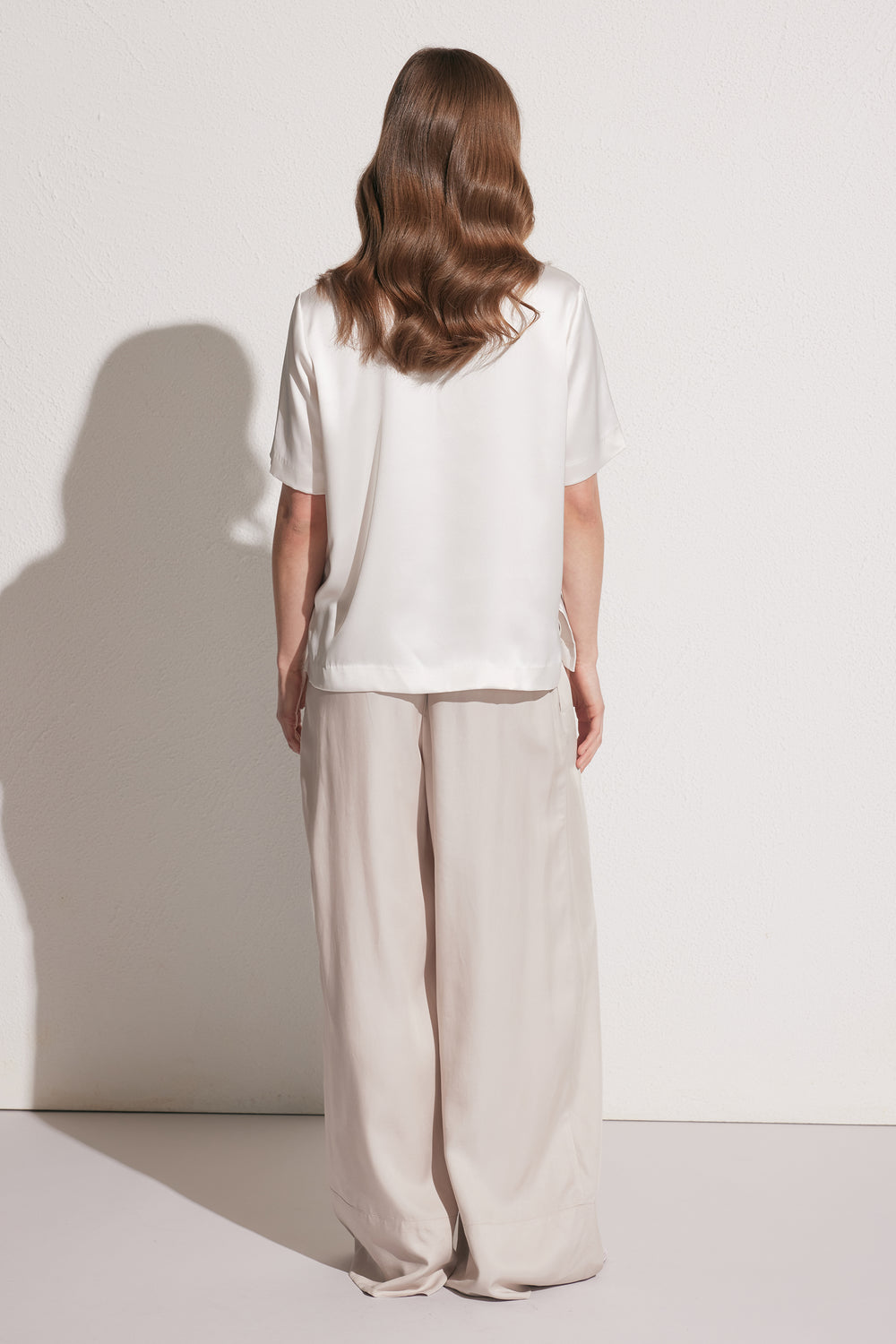 Back view of woman wearing loose beige wide-leg pants with a white short-sleeve satin tee, against a plain wall