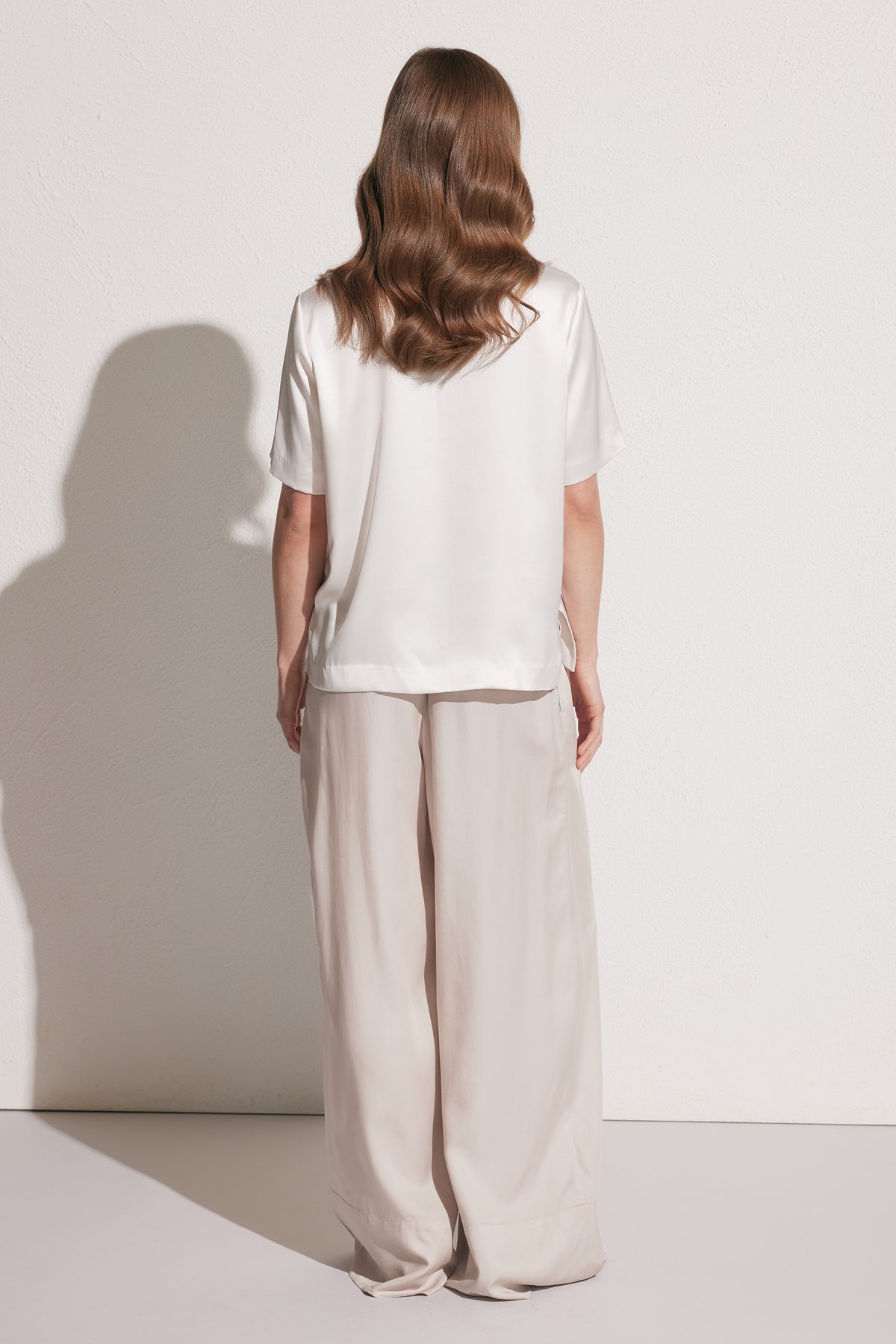 Back view of woman wearing loose beige wide-leg pants with a white short-sleeve satin tee, against a plain wall