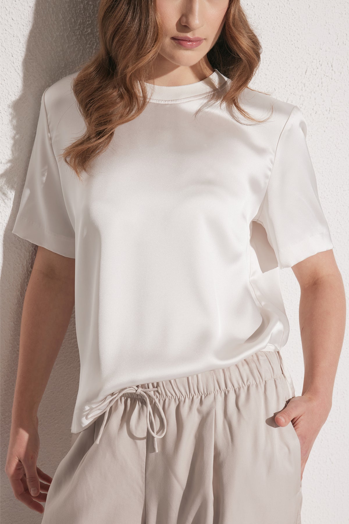 Woman wearing a smooth white satin tee with short sleeves paired with light beige drawstring pants against a textured wall