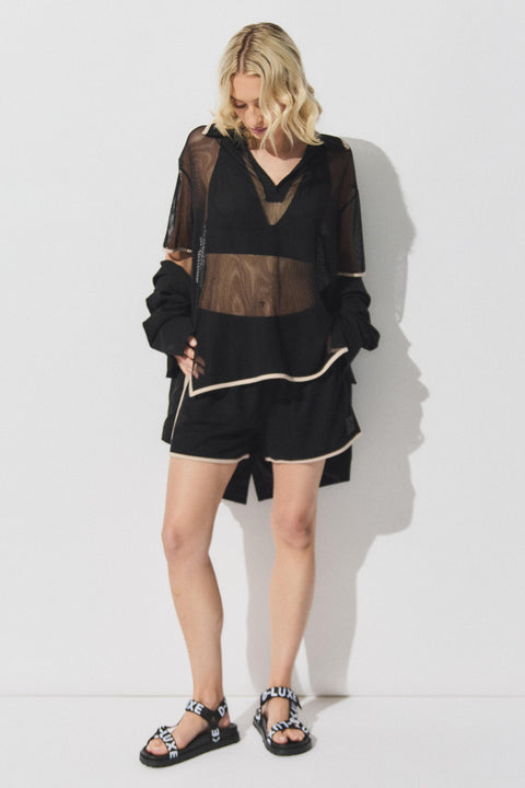 Woman wearing black knit shorts with beige trim and a sheer black top layered over a black bralette and sandals