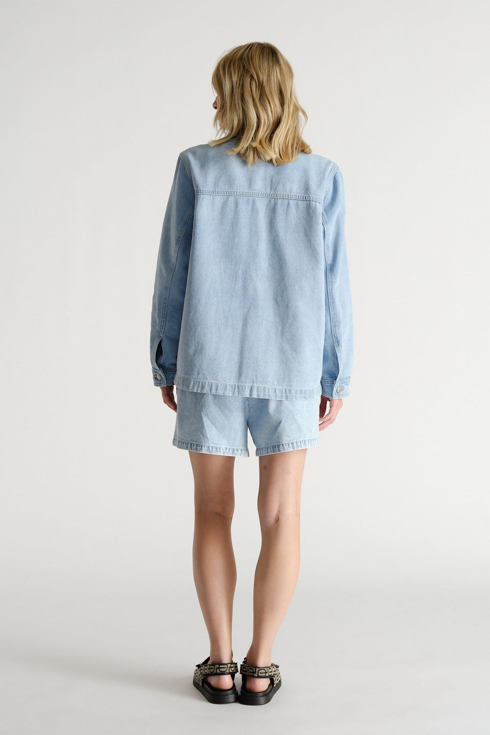 Woman seen from behind wearing a light blue denim jacket and matching D-Luxe shorts with black patterned sandals