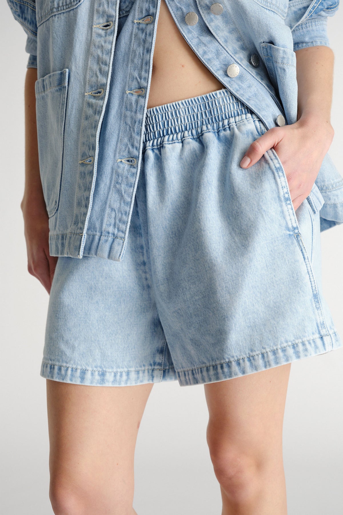 Close-up of light wash denim pull-on shorts with elastic waistband paired with a matching oversized denim jacket