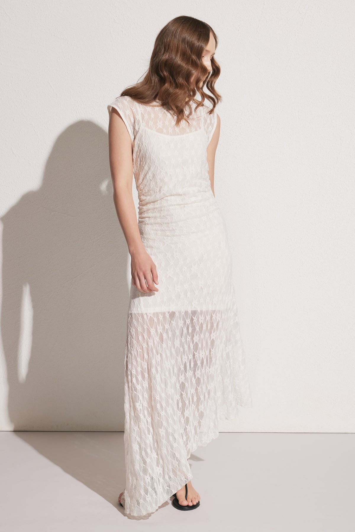 Woman wearing a cream lace asymmetric dress with cap sleeves, styled with black flat sandals, standing against white backdrop