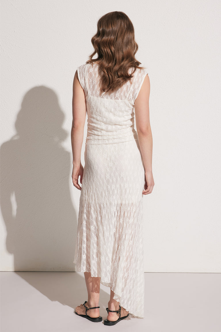 Woman wearing a cream lace dress with an asymmetric hem and ruched details, paired with black flat sandals, back view