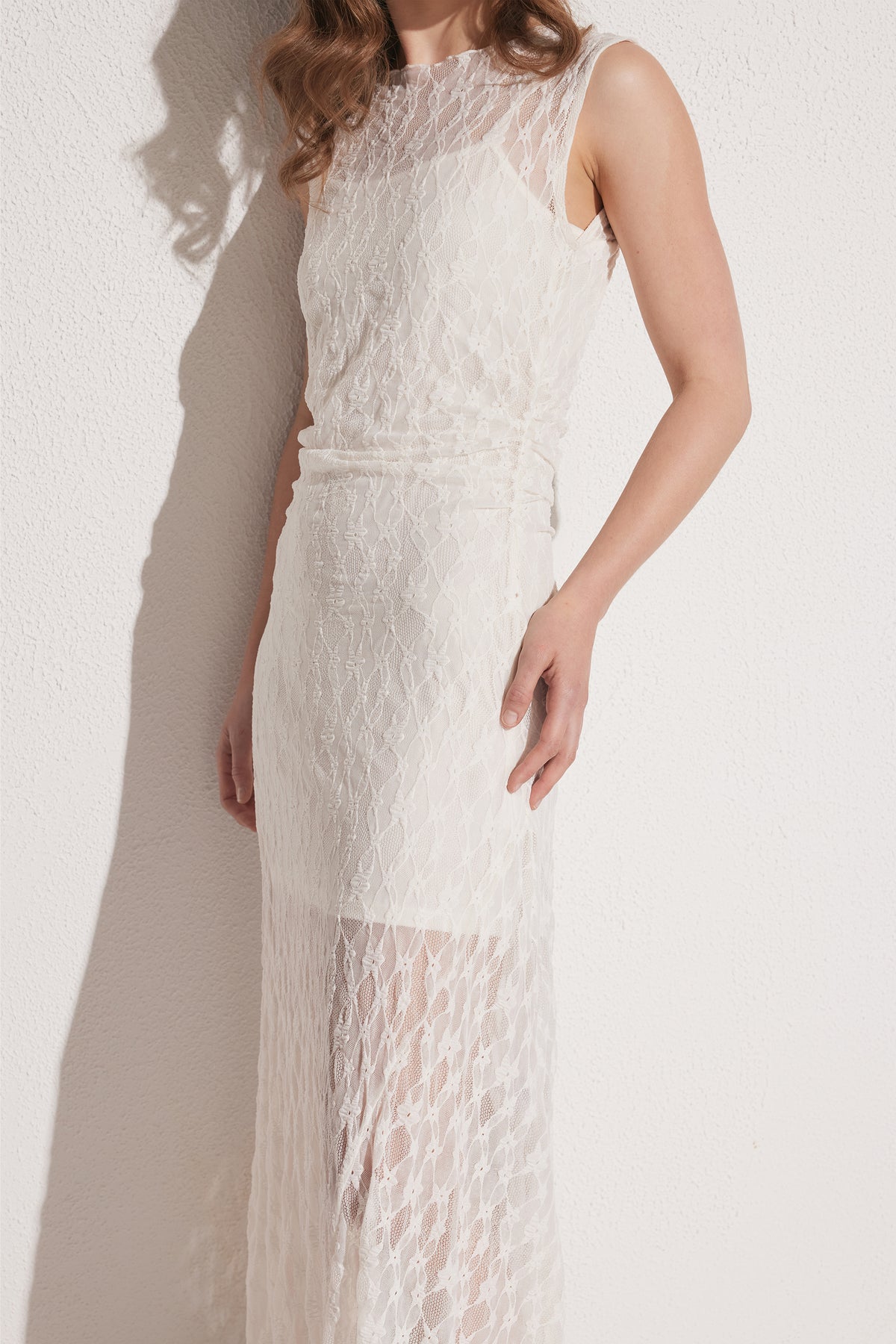 Woman wearing a sleeveless ivory lace dress with an asymmetric silhouette, standing against a textured wall.