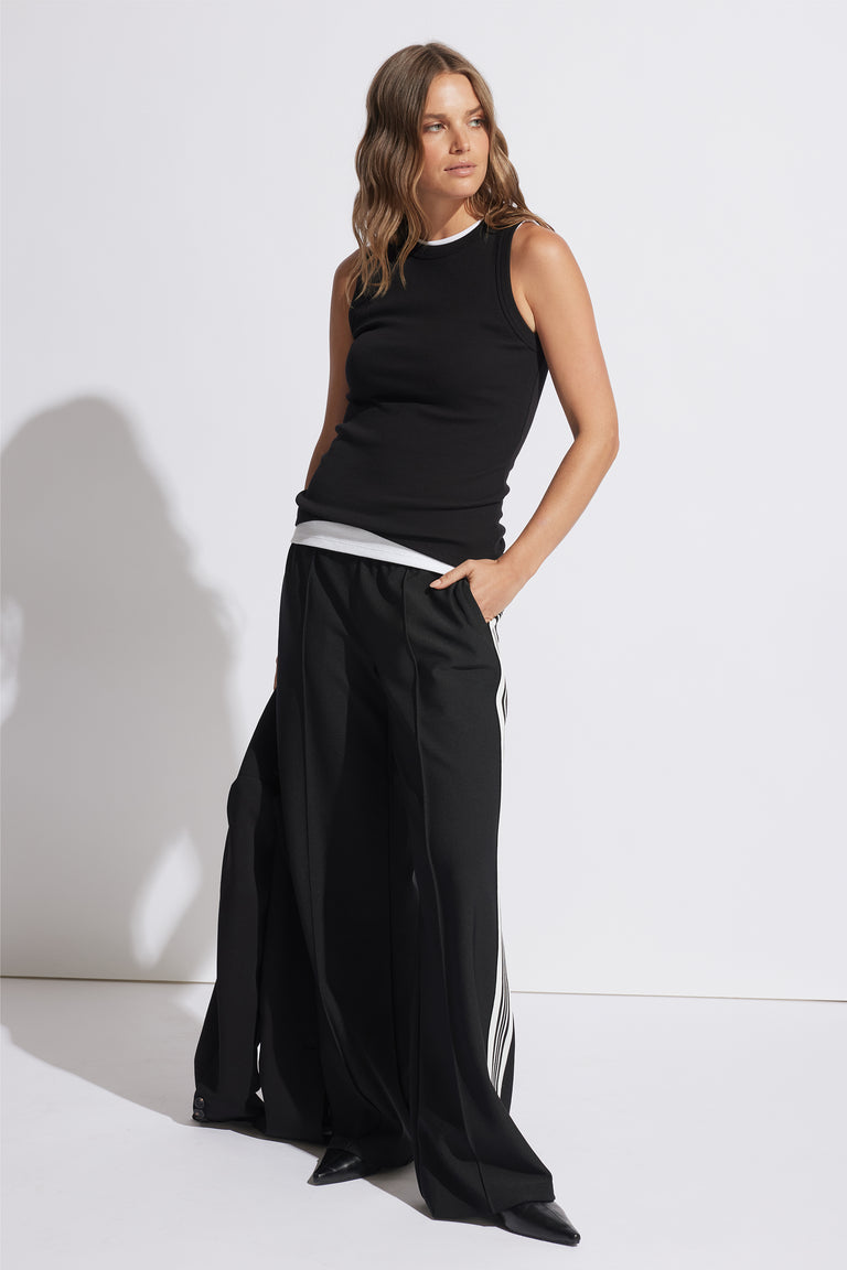 Woman wearing a fitted black ribbed tank with wide-leg black pants featuring white side stripes in a minimalist studio setting